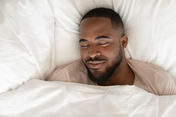 Top view of happy african American man sleeping in comfortable white bed seeing good pleasant dreams, calm biracial male feel fatigue resting napping in cozy bedroom under linen bedding sheets